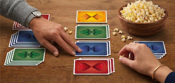 Two players laying colorful SCORE 4 cards onto shared piles on a wooden table, with a bowl of popcorn